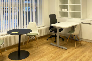 Behandlungszimmer und Beratungsbereich einer modernen Gynäkologie-Praxis mit hellem Vinylboden Expona von Objectfloor in Holzoptik. Zu sehen sind ergonomische Stühle, ein runder Tisch und ein höhenverstellbarer Schreibtisch vor einem grossen Fenster mit Jalousien.